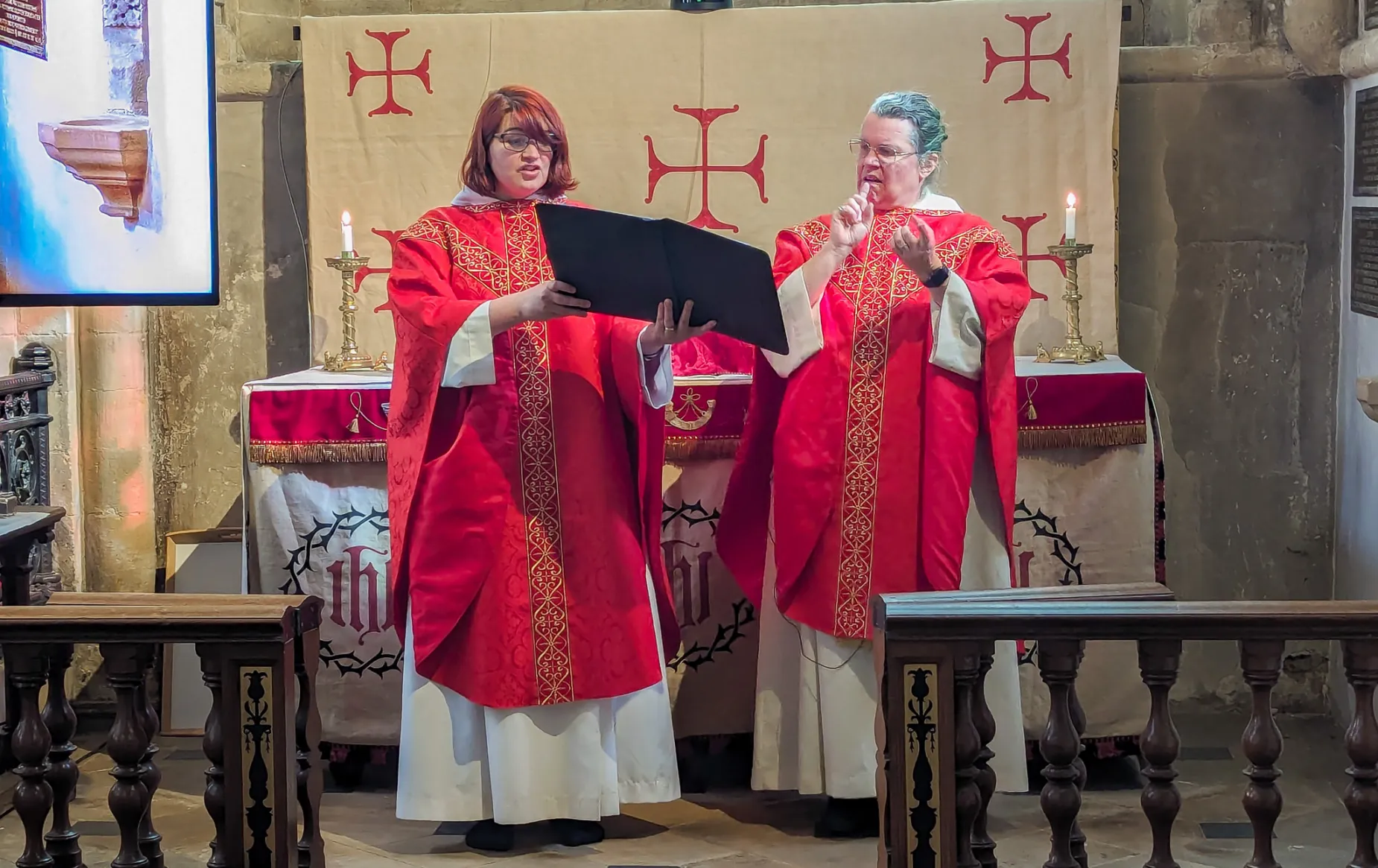 Revd Philippa White and Reverend Hannah Lewis celebrate the eucharist together. Revd Philippa is holding the book of the liturgy, while Revd Hannah is reciting the liturgy in British Sign Language, her hands in mid air. They are both wearing red chausibles, appropriate to the final weeks of Lent, and standing in front of the Remembrance Chapel altar.