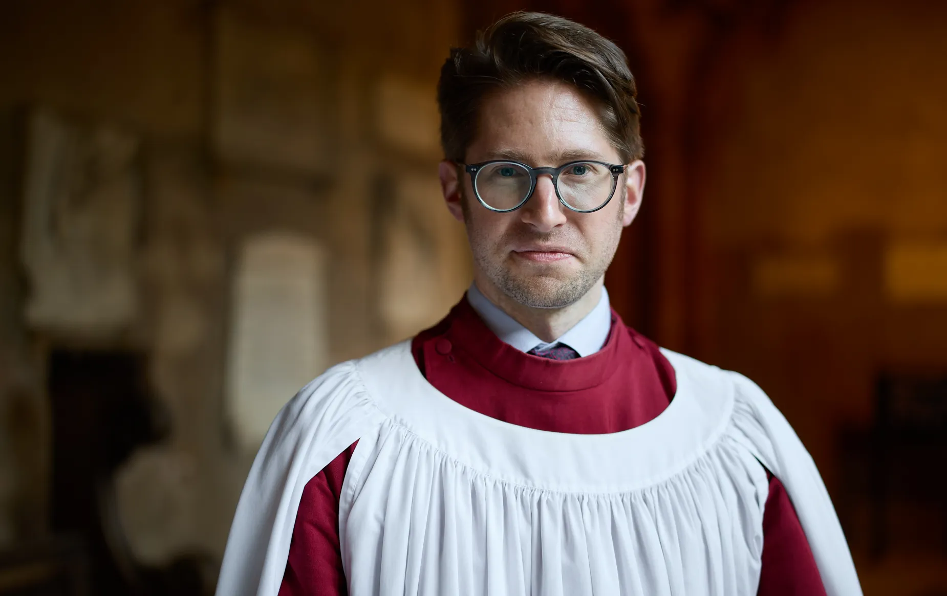A portrait of organist Peter Holder in liturgical clothing, standing in the cloisters.