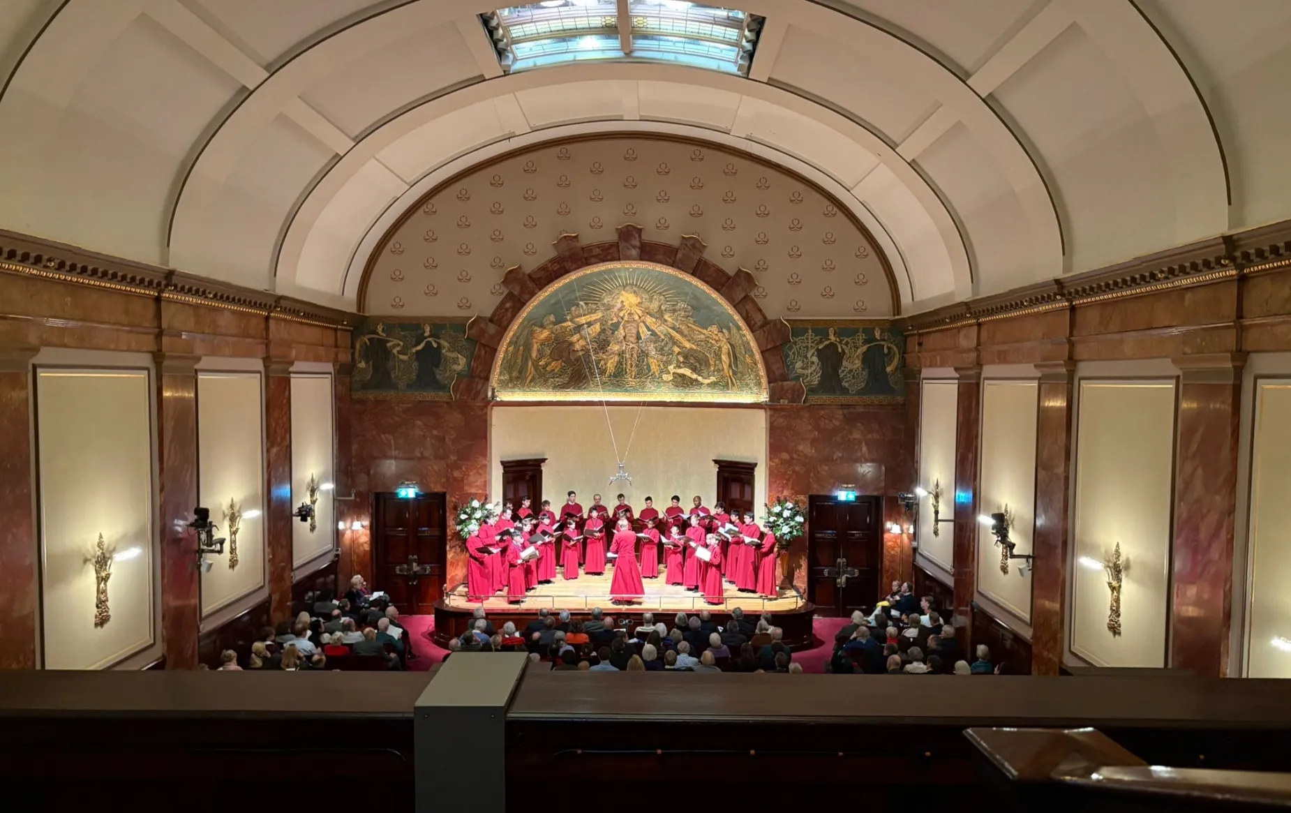 The cathedral choir, in robes, perform in a full wigmore hall, a grand chamber music venue in London with a neoclassical appearance