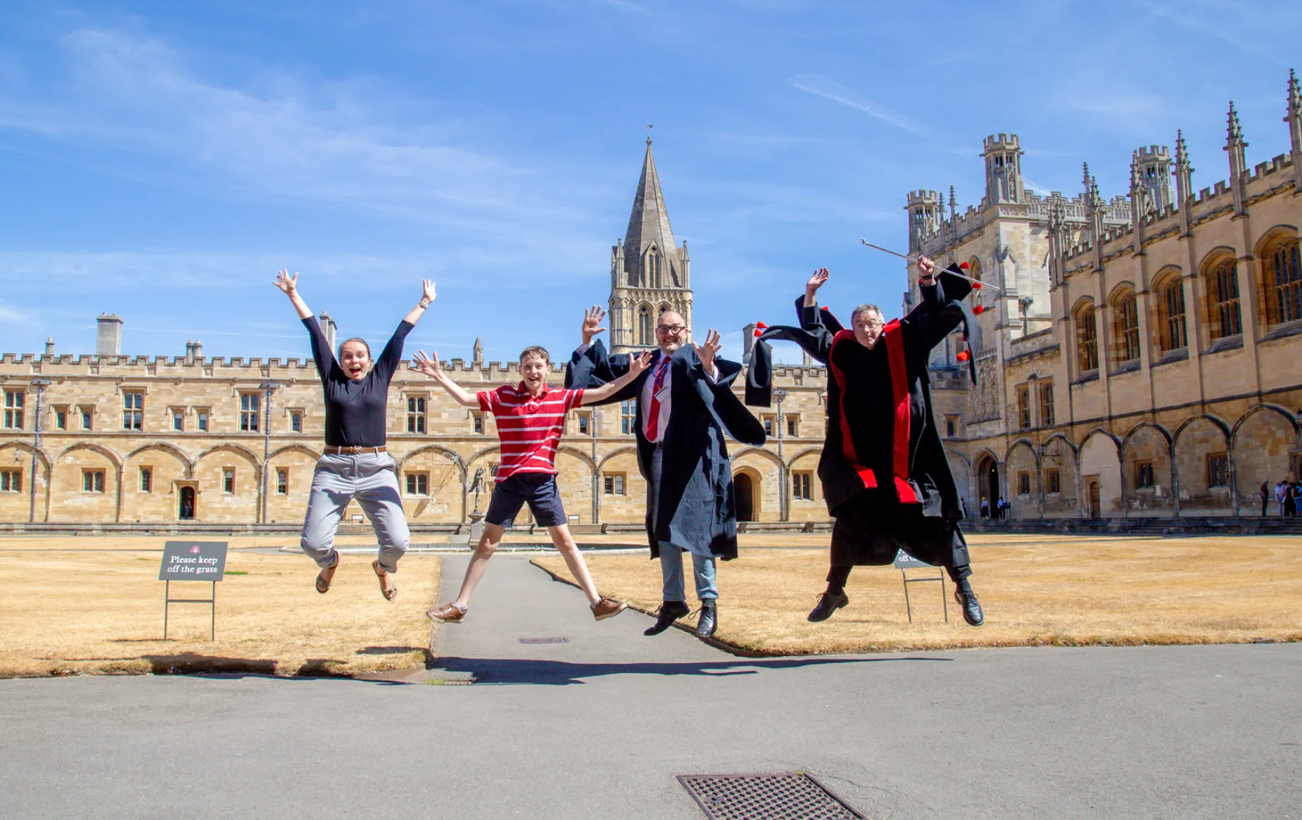 Jackson Brown jumps for joy in tom quad with teh cathedral spire behind him, joined by the College Chaplain, Cathedral operations manager in academic gown, and head verger in his red and black vergers gown.