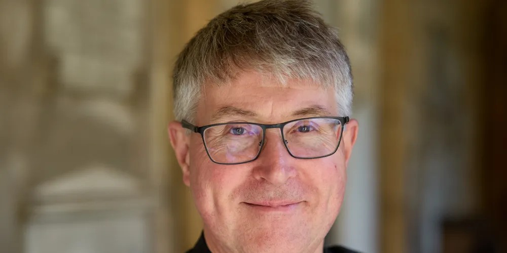 Portrait of The Revd Canon Peter Moger, Sub Dean, in clerical collar and academic gown, standing in the Cathedral Cloister