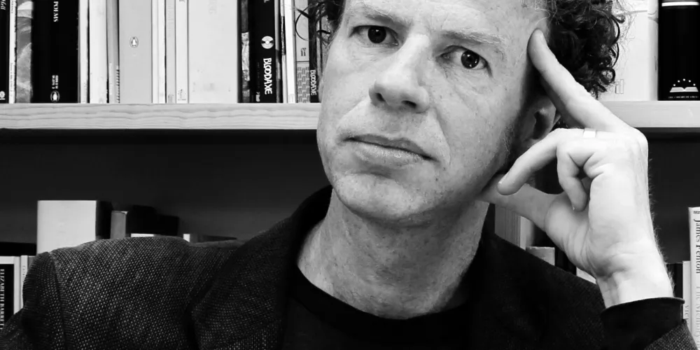 Black and white photo of Matthew Hollis, a man with short curly hair in front of bookshelves