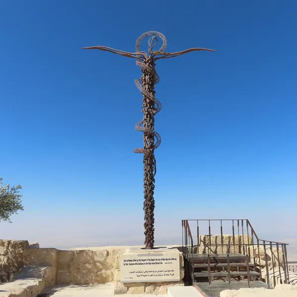 A monument commemorating the story of the Israelites in the desert being saved by 'the brazen snake', consisting of a stylized tall bronze cross with a snake curled around it. It stands on a hill by a 4th century Byzantine church and overlooks the negev desert under a deep blue sky.