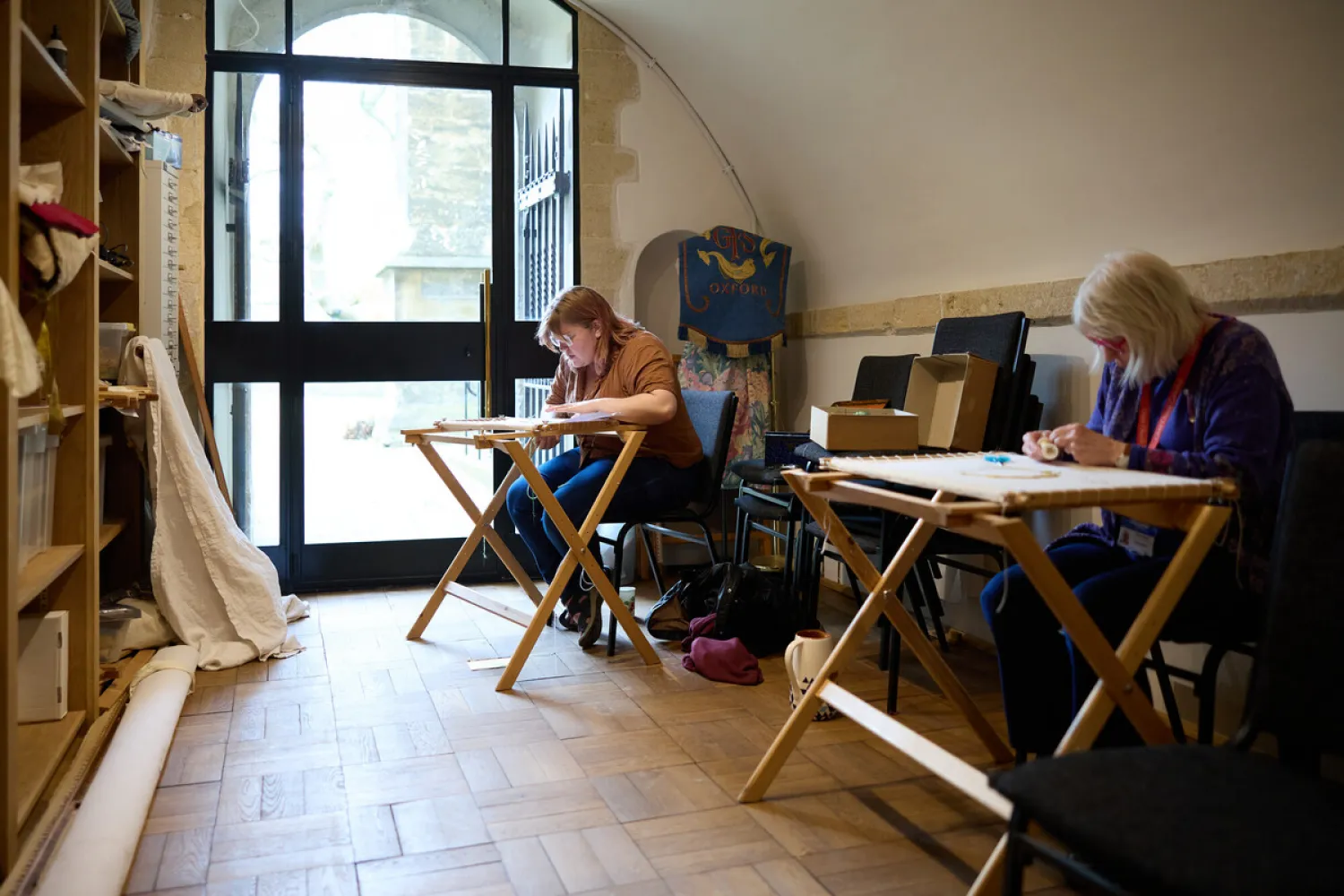 Two embroiderers are working at folding wooden embroidery frames in the cathedral Slype, a small room with a wooden floor and a sloping ceiling. The Slype is also used by the adult singers of the Choir and so there are shelves on the left with some robes and music. Light is streaming into and filling the room through a large windowed door opening to the garden beyond.