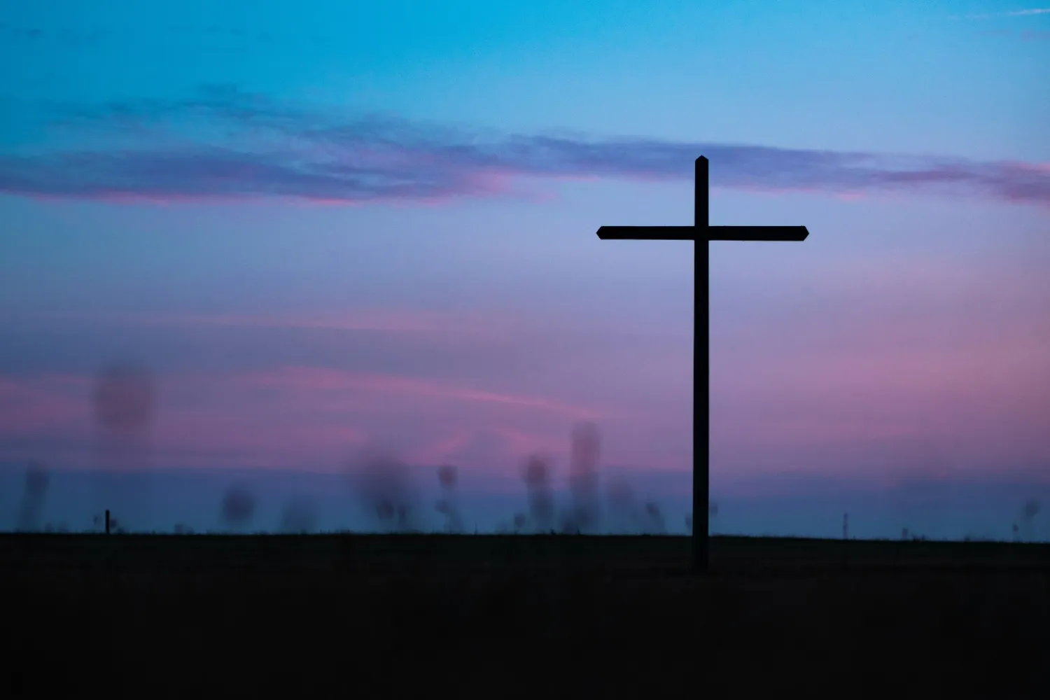 A distant Christian cross stands silhouetted against a darkening blue and pink sky in the final stages of twilight