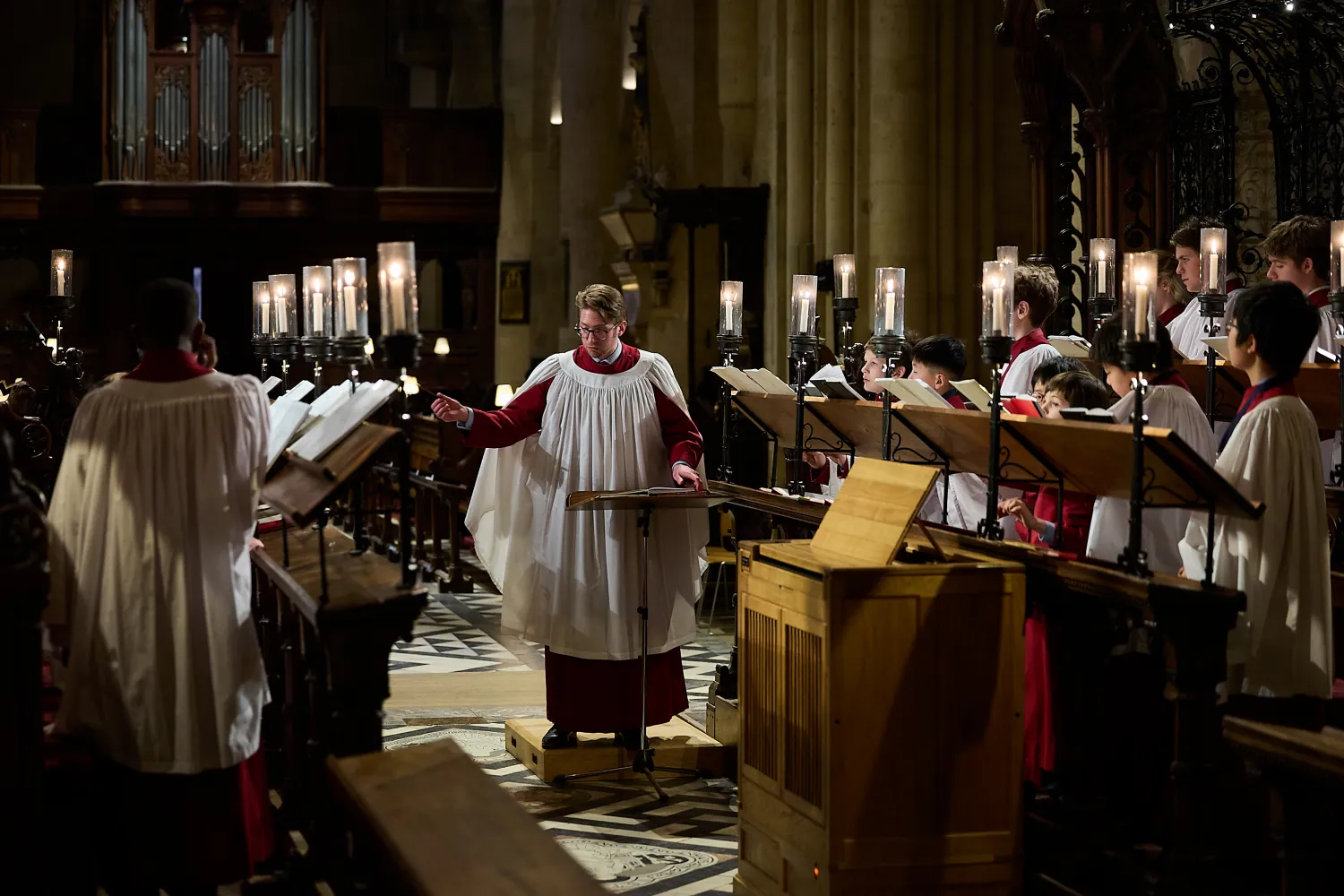 Peter holder conducts the Cathedral choir, who are wearing their liturgical clothing. They are in the choir stalls, lit by candles.