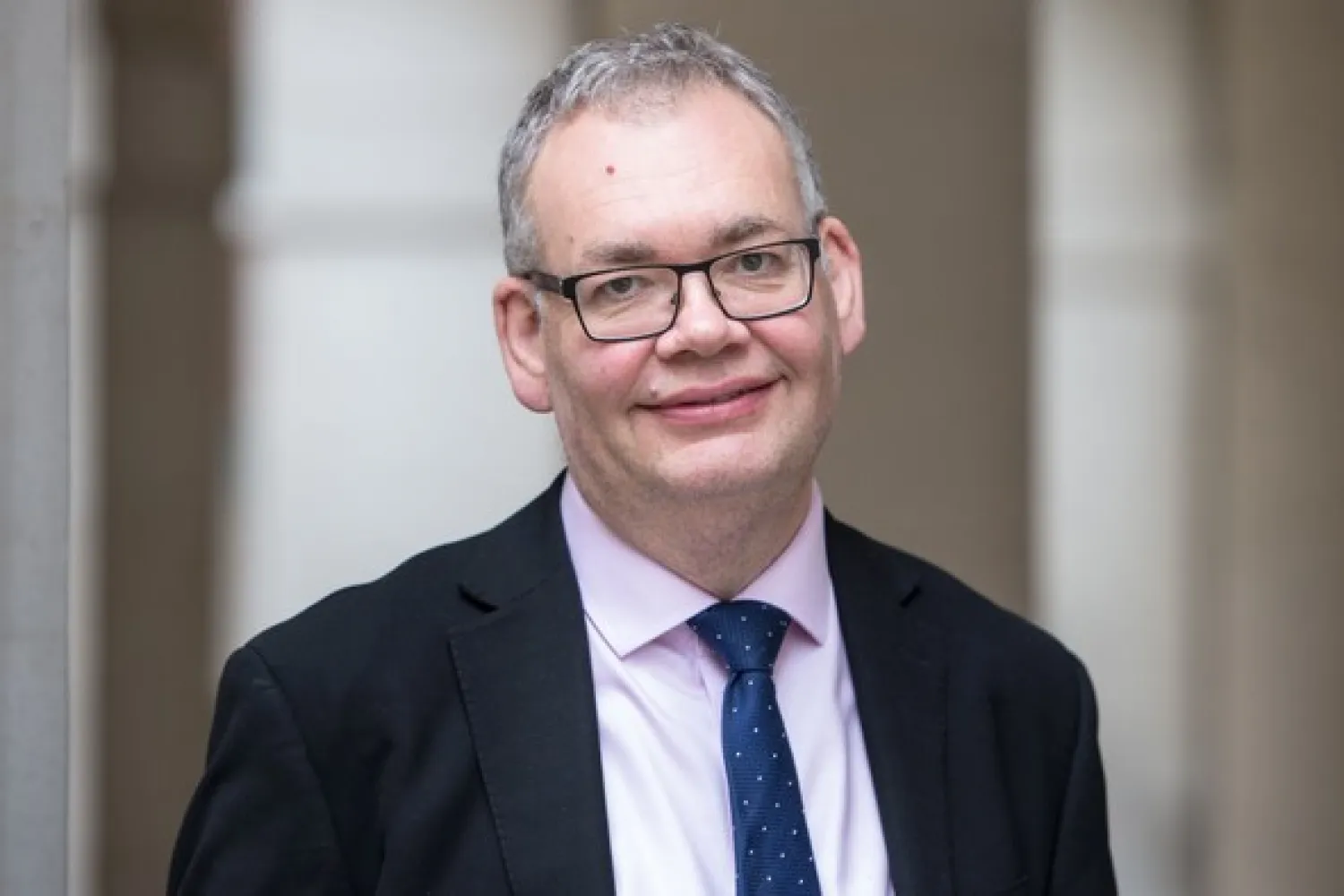 Roger sayer, in a dark suit and blue tie, with stone colonades in the background.