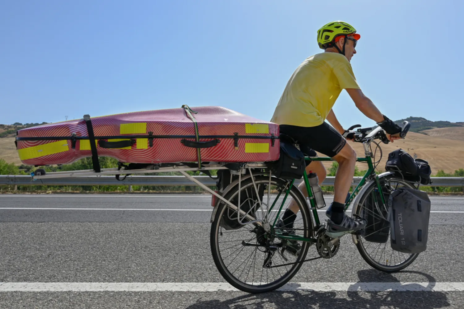 Kenneth Wilson rides a bike with a cello strapped on the back on a hot sunny day along a road.
