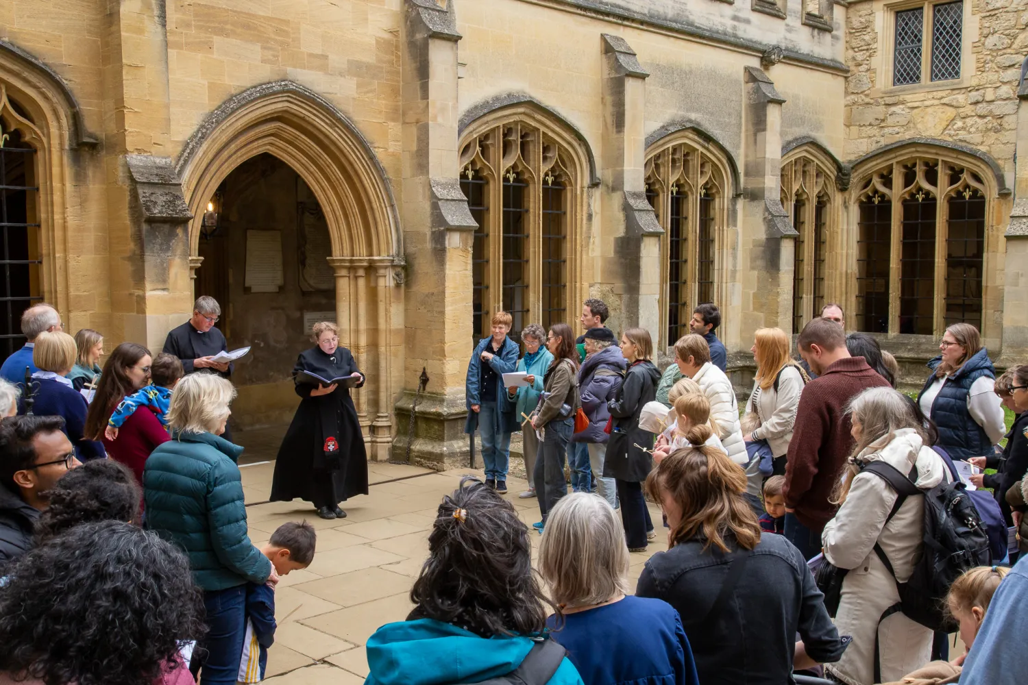 A group of children and their guardians stand in the middle of the cloisters as a robed verger reads to them during our Journey to bethlehem service in 2025