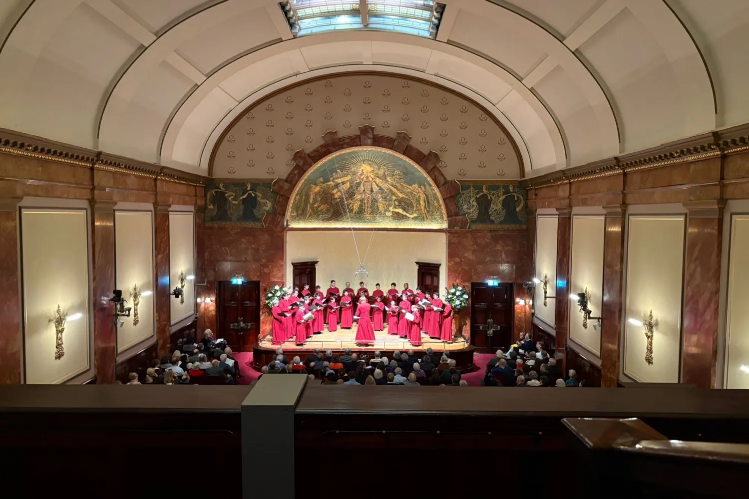 The cathedral choir, in robes, perform in a full wigmore hall, a grand chamber music venue in London with a neoclassical appearance