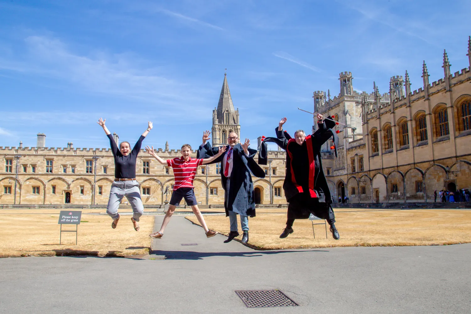 Jackson Brown jumps for joy in tom quad with teh cathedral spire behind him, joined by the College Chaplain, Cathedral operations manager in academic gown, and head verger in his red and black vergers gown.