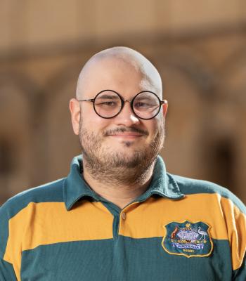 Portrait of Davide Sutto smiling at the camera, wearing round black glasses and a green and gold Australian rugby shirt. He stands against a softly blurred, warm-toned background in natural sunlight.