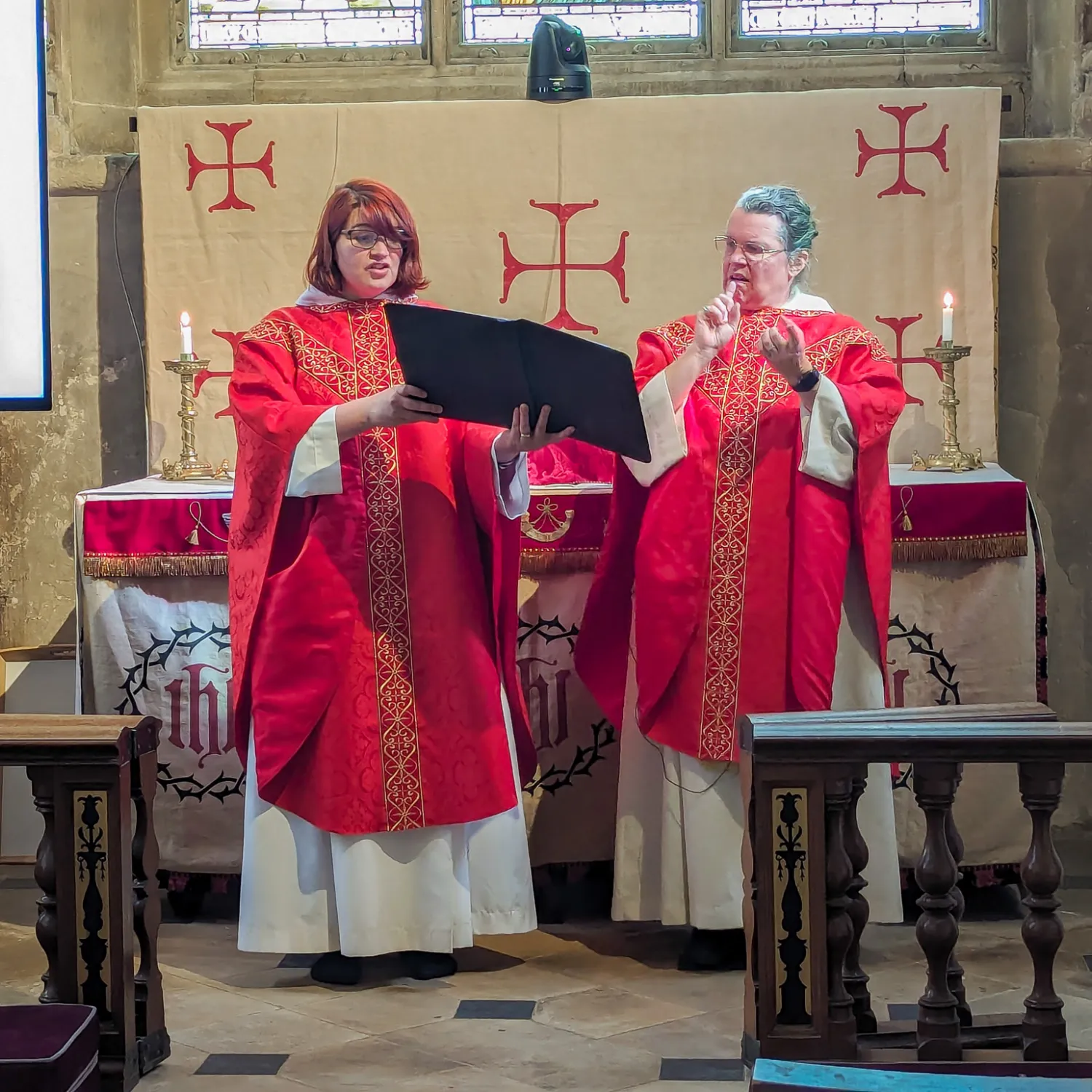 Revd Philippa White and Reverend Hannah Lewis celebrate the eucharist together. Revd Philippa is holding the book of the liturgy, while Revd Hannah is reciting the liturgy in British Sign Language, her hands in mid air. They are both wearing red chausibles, appropriate to the final weeks of Lent, and standing in front of the Remembrance Chapel altar.