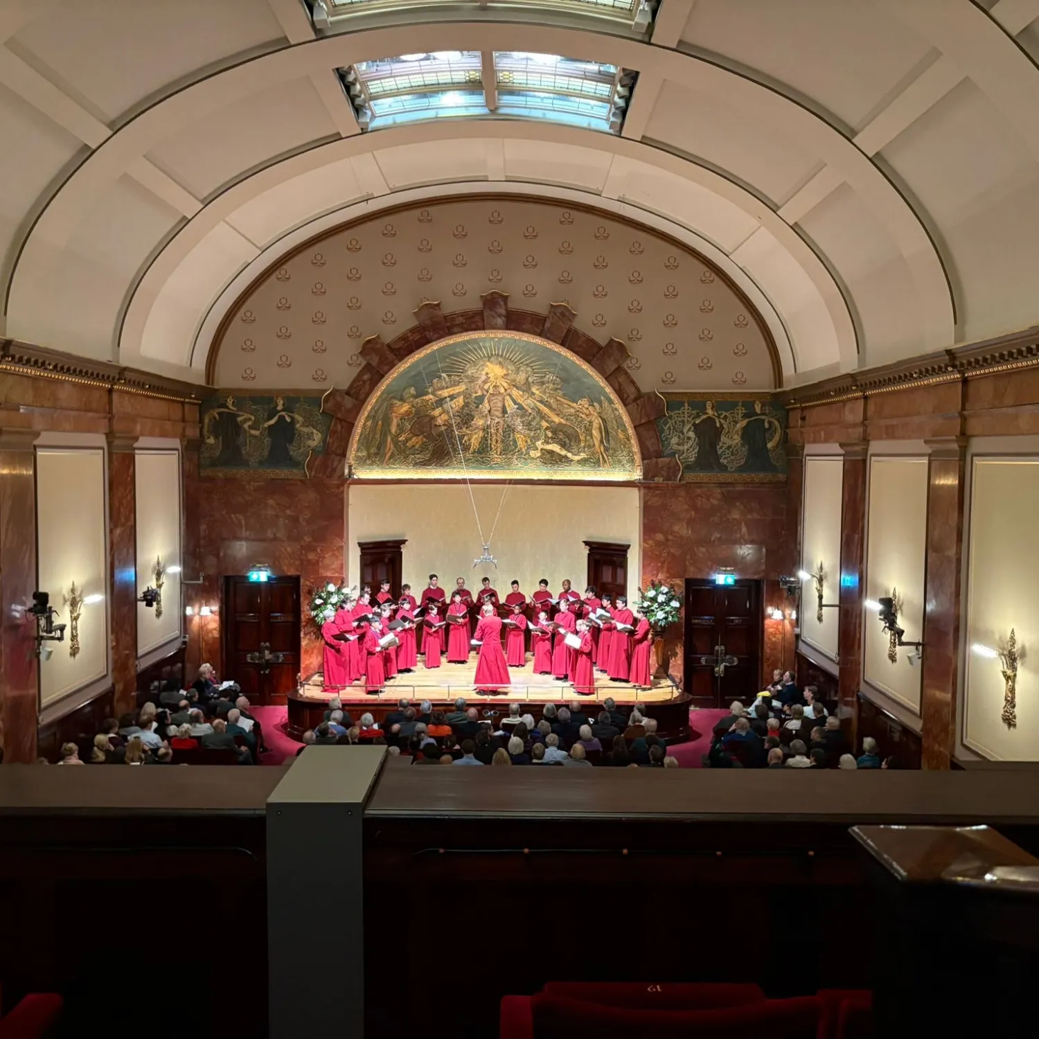 The cathedral choir, in robes, perform in a full wigmore hall, a grand chamber music venue in London with a neoclassical appearance