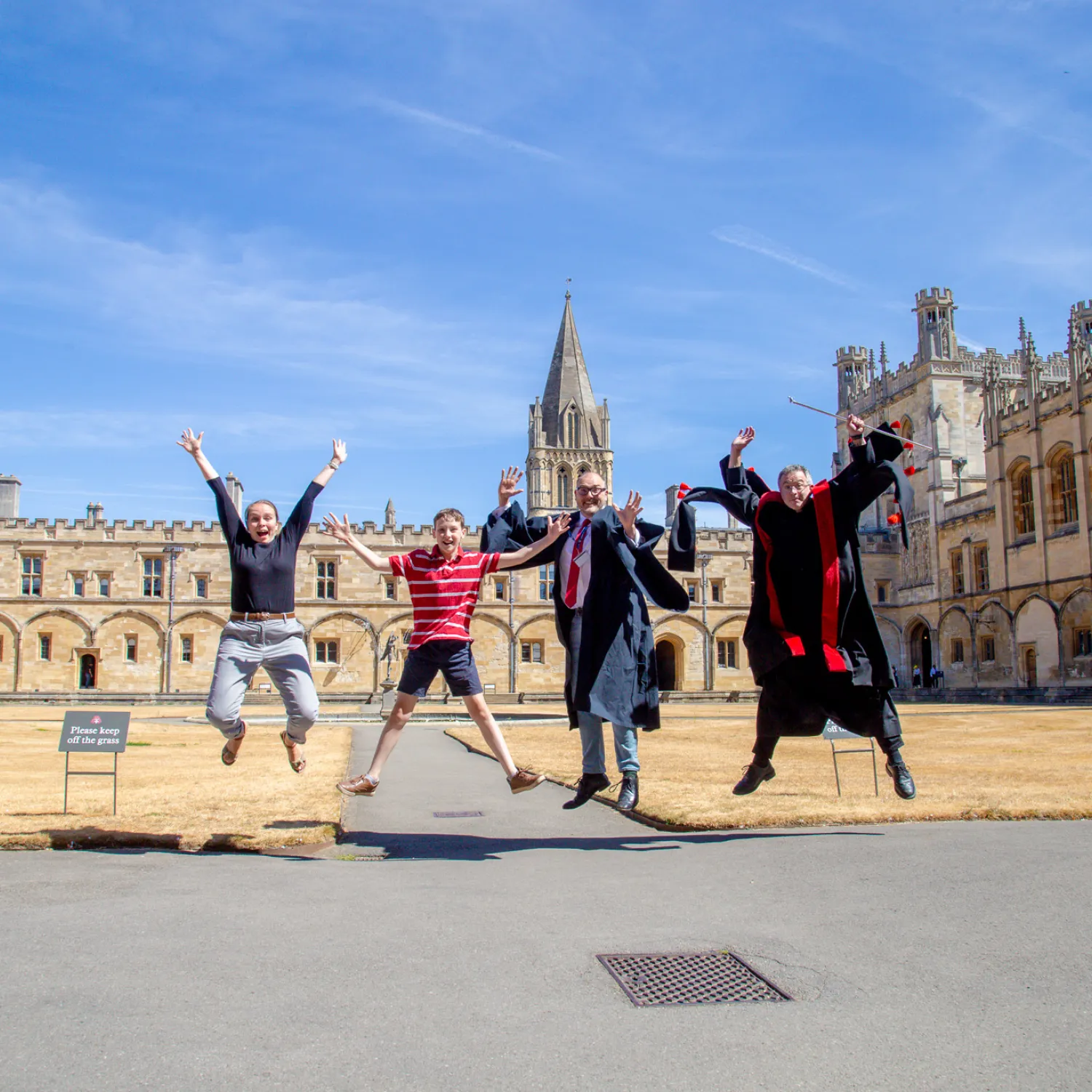 Jackson Brown jumps for joy in tom quad with teh cathedral spire behind him, joined by the College Chaplain, Cathedral operations manager in academic gown, and head verger in his red and black vergers gown.